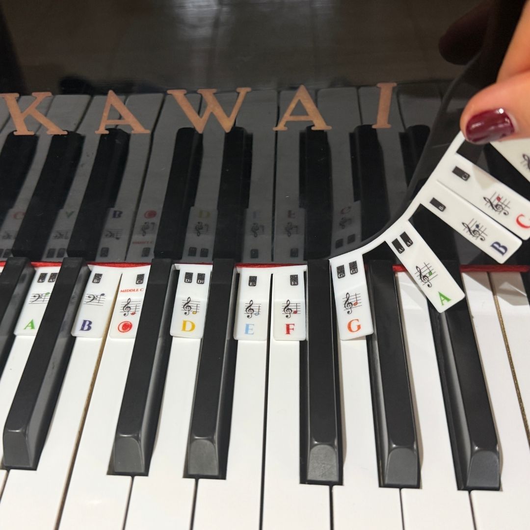 A close-up of a piano keyboard with small white instructional cards placed on the keys, and a hand holding a stack of similar cards. The word KAWAI is visible above the keys, indicating the Kawai brand.